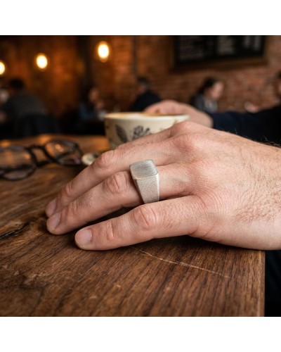 Bague rectangulaire argentée portée sur le doigt d'une main posée sur une table en bois.