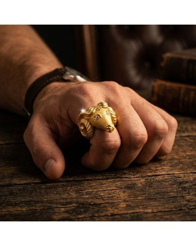 Anneau doré en forme de tête de bélier porté sur le doigt, posé sur une surface en bois; motifs gravés et brillance discrète. Ba