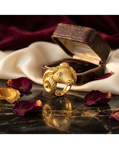 Bague dorée en forme de tête de bélier près d’un coffret ancien et pétales séchés sur marbre, reflet visible. Mot-clé: Bague BEL