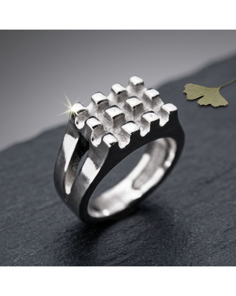 Bague GAUFFRE argentée, ring moderne à motif grille 3D brillant sur une surface sombre, avec fleur fanée en arrière-plan