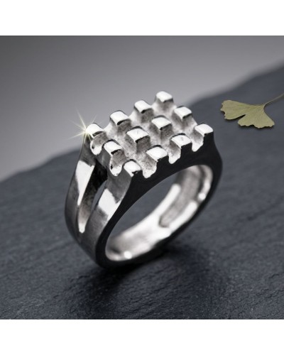 Bague GAUFFRE argentée, ring moderne à motif grille 3D brillant sur une surface sombre, avec fleur fanée en arrière-plan
