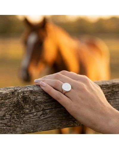 Bague rond de longe en argent rhodié gravée du logo Murial portée sur le doigt, cadre bois et cheval au fond.
