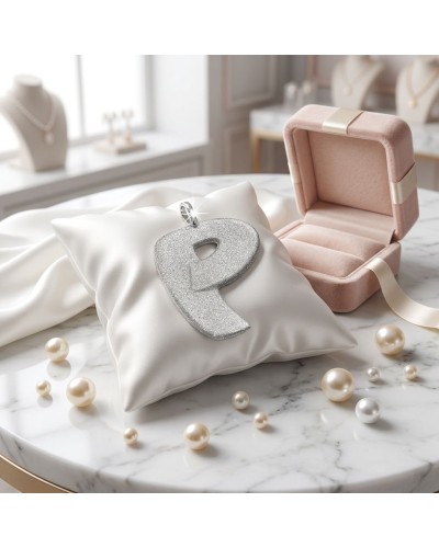 Pendentif en forme de lettre P argentée étincelante posé sur un coussin blanc, avec boîte rose et perles autour. Chaîne offerte.