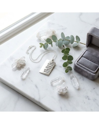 Pendentif KEKAYAAN argenté avec chaîne, posé sur plateau marbre près d’éléments en pierre et une tige de eucalyptus, boîte à bij