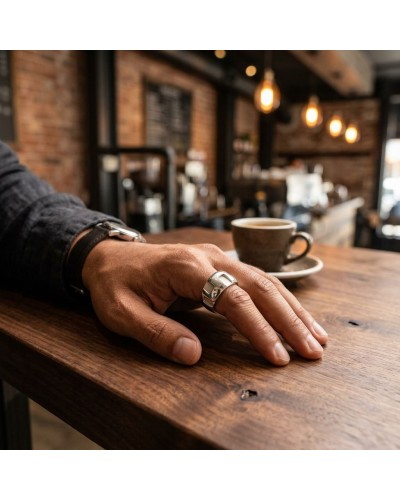 Bague GREC en métal argenté portée sur le doigt d’une main posé sur une table en bois, café en arrière-plan.