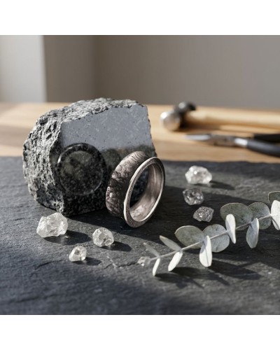 Bague ANNEAU SERRE en métal sombre avec motif relief, posée sur pierre noire, entourée de petits cristaux et feuilles d’eucalypt