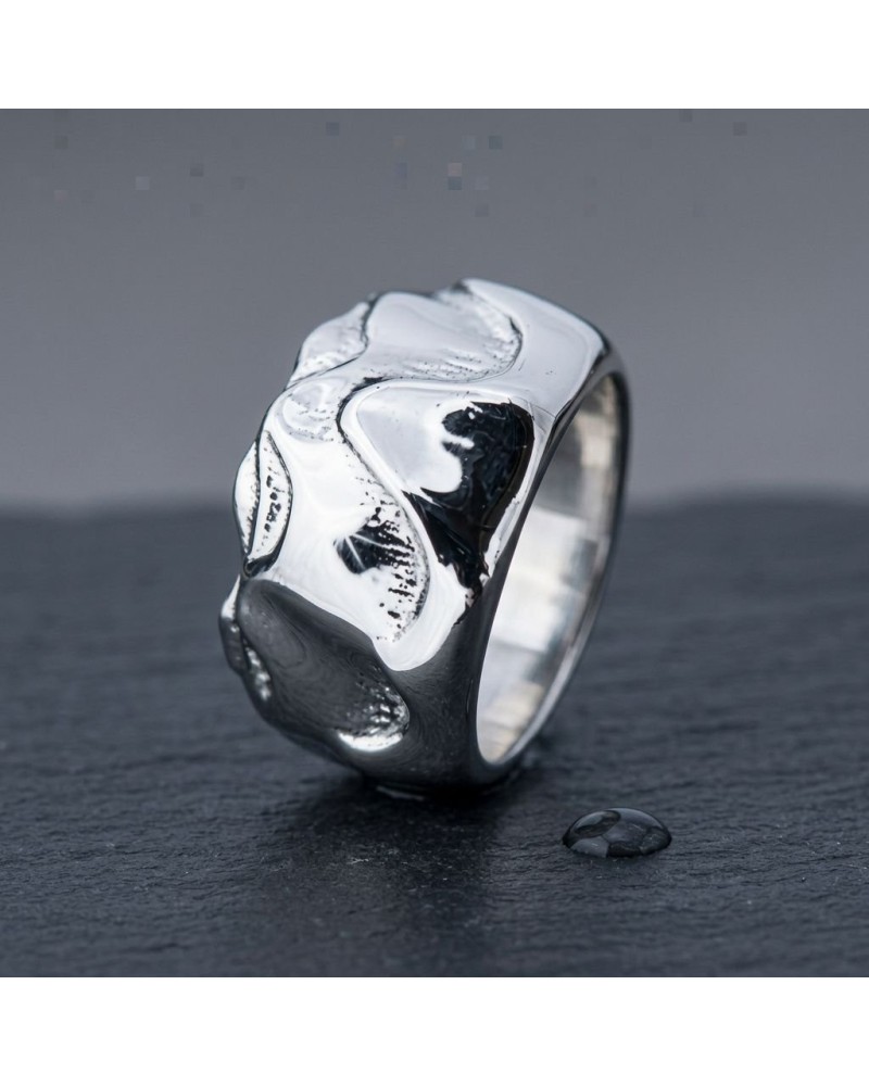 Bague TEMPÊTE en argent poli avec motifs ondulants, posée sur une surface noire, petite pierre noire à côté.