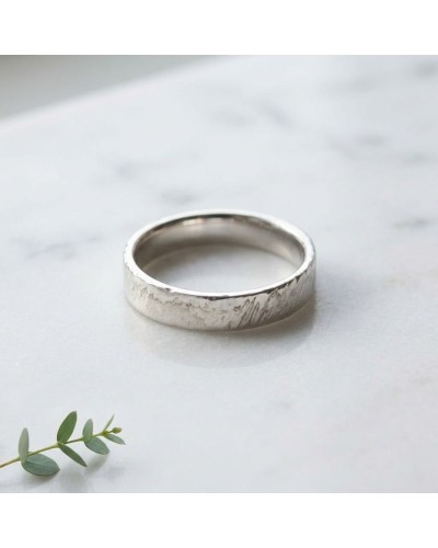 Alliance homme argentée au motif strié, ring plat posé sur marbre, branche verte au premier plan.