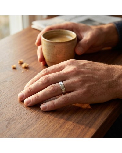 Alliance effet strié pour homme en or: petite bague argentée uni-forme sur doigt masculin, posé sur table en bois avec tasse et