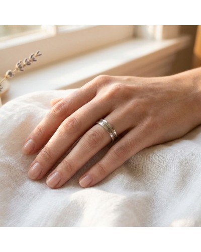 Alliance PEACE femme en or portée sur l’annulaire, main reposant sur tissu blanc près d’une fenêtre.