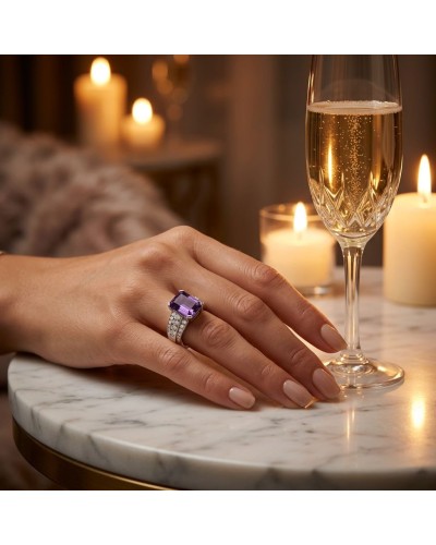 Bague NINA élégante avec pierre violette sur doigt posé sur table en marbre, champagne flûte et bougies en arrière-plan.
