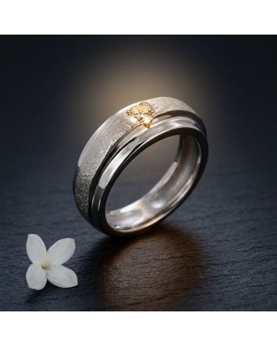 Alliance homme en or sertie d’un diamant doré, sur fond sombre avec fleur blanche à côté.