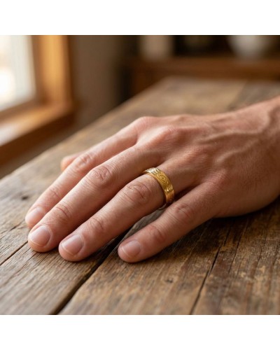 Alliance homme en or gravée portée sur l'annulaire d'une main masculine, posé sur une table en bois.