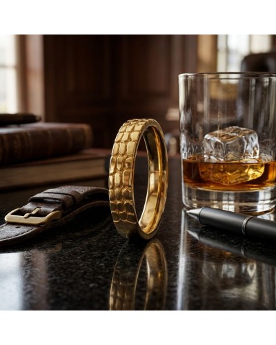 Alliance demi-effet crocodile en or posée sur une surface sombre, entourée d’un verre de whiskey et d’un bracelet en cuir.