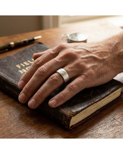 Anneau argenté large porté sur la main posée sur un livre ancien, outils et montre flous en arrière-plan. Bague ANNEAU.