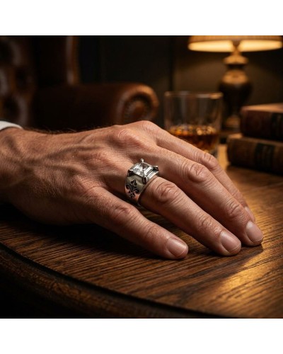 Bague argentée masculine épaisse avec motif fleur de lis portée sur le annulaire, posé sur une table en bois près d’un verre et